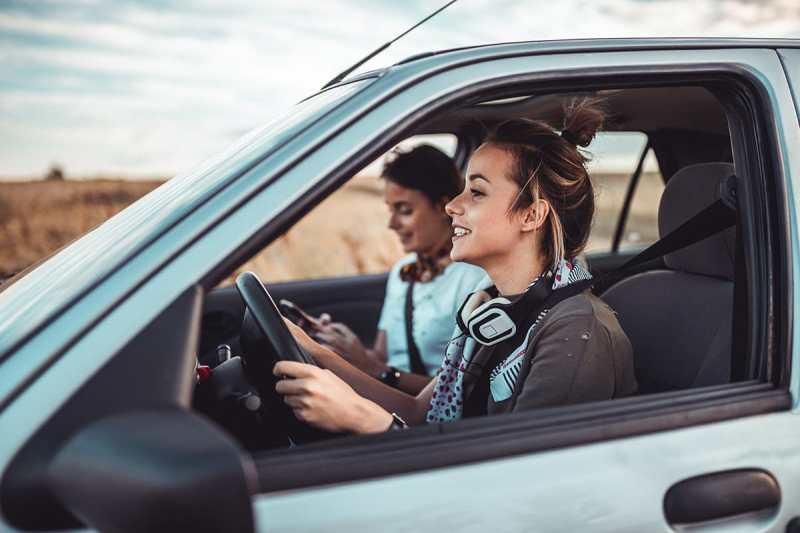 Two friends enjoying a fun car ride, with one driving and the other engaged with her phone