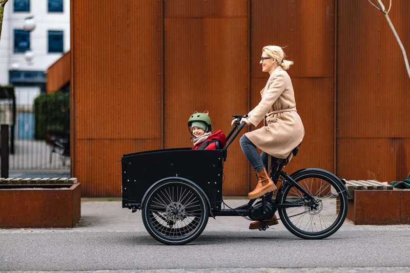 Woman riding a cargo bicycle with a child, promoting family-friendly outdoor transportation