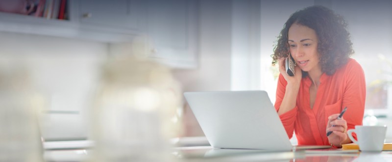 A woman on the phone while working on a laptop at home, engaged in a conversation or task