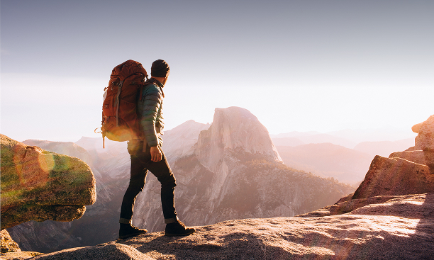 Hiker with a backpack standing on a rock outcrop, gazing at a distant mountain landscape during sunset