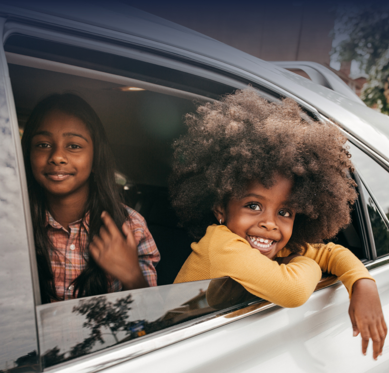 Two girls happily enjoying a car ride, one smiling out the window, while the other looks on playfully