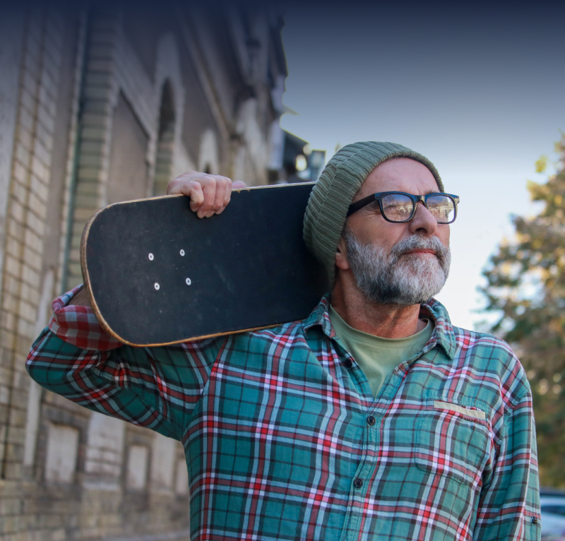 Older man holding a skateboard, reflecting on city life in an urban setting