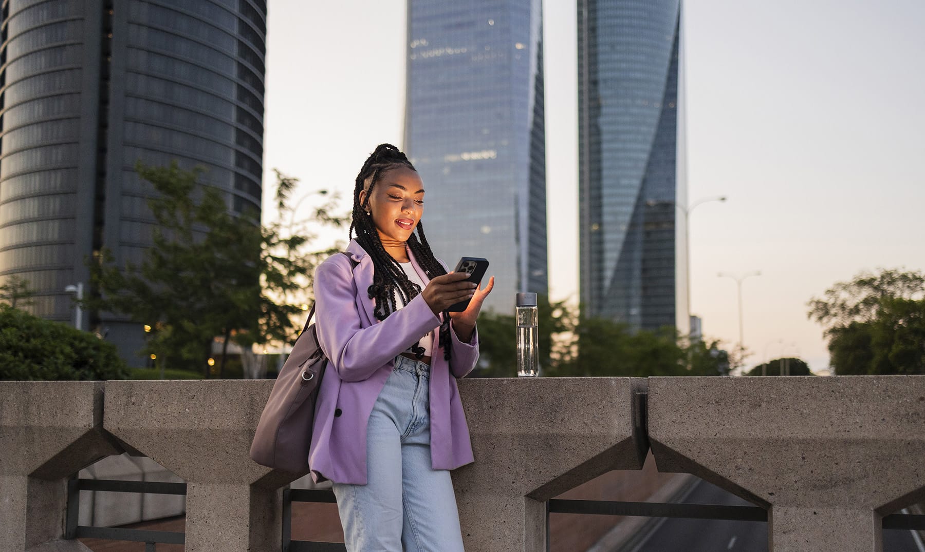 Woman using her phone while standing by a concrete barrier in an urban setting during sunset