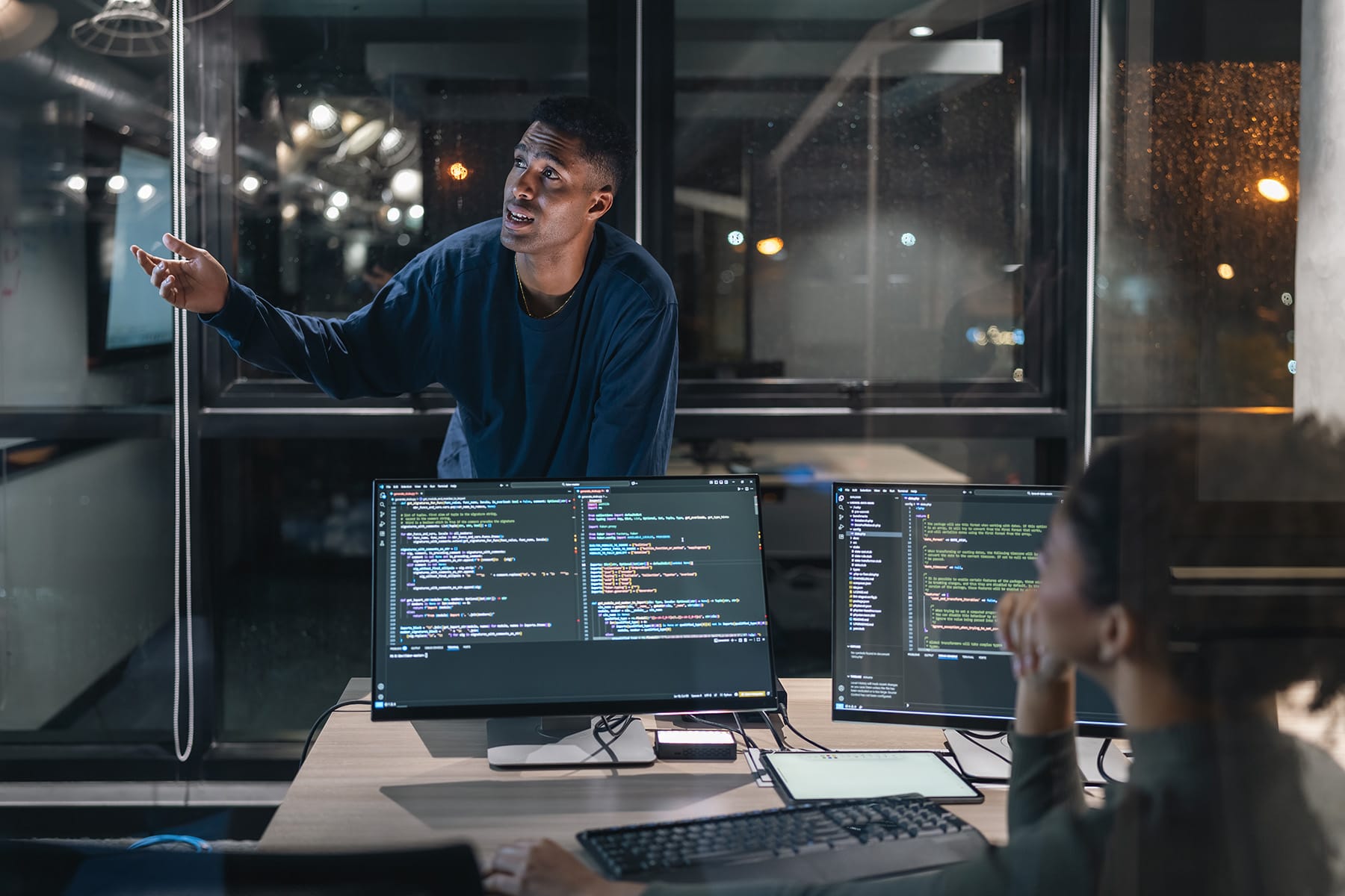 A team member presents technical information during a meeting in a modern office with multiple computer screens