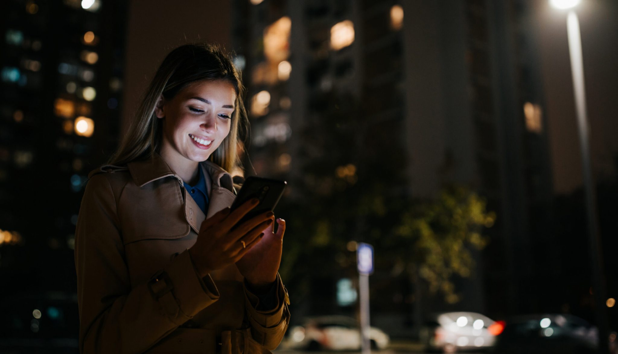 A woman smiles while using her phone outdoors at night near buildings