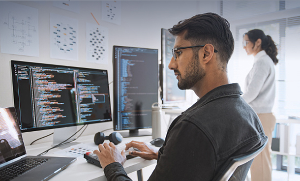 A programmer working at a desk with multiple monitors while a colleague observes in the background